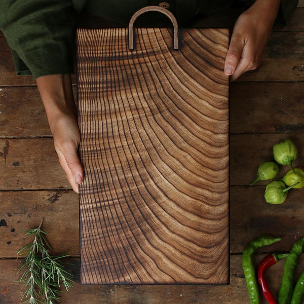 Handmade by Ambrose Vevers. These chopping boards are made from ash wood, which has been scorched with a naked flame and finished with food safe oil. A leather loop is at the top of the board for easy display and storage. The shape of this board is a rectangle.