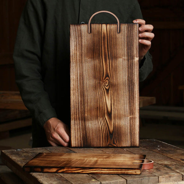 Handmade by Ambrose Vevers. These chopping boards are made from ash wood, which has been scorched with a naked flame and finished with food safe oil. A leather loop is at the top of the board for easy display and storage. The shape of this board is a rectangle.