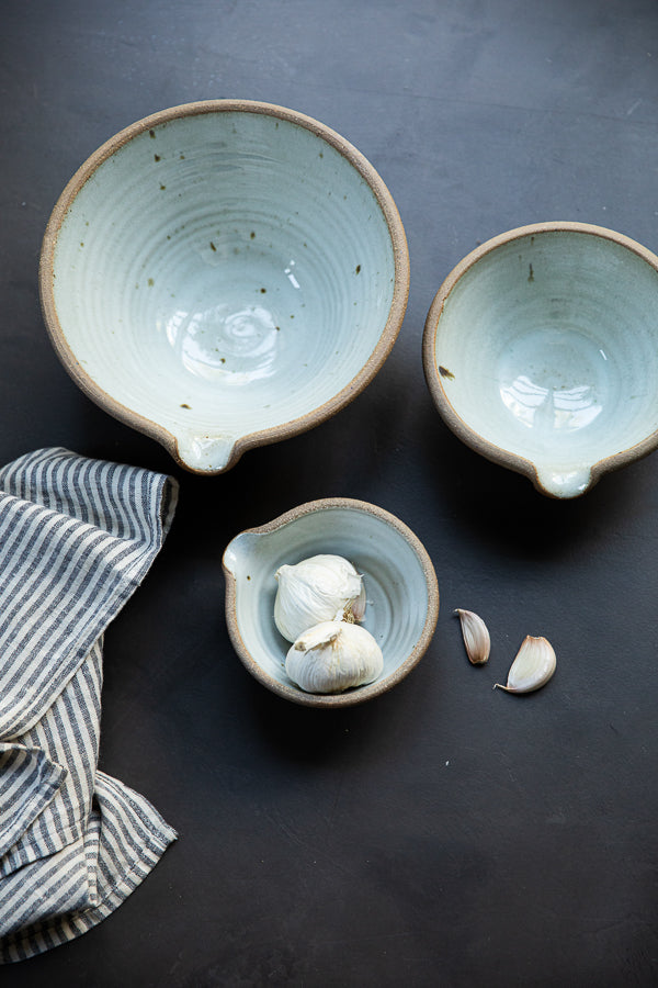 Handmade by the Leach Pottery Studio in St Ives. A set of three mixing bowls each finished with a lip for pouring. These bowls are glazed on the inside with a very pale duck egg blue dolomite glaze. The outside is unglazed for a natural and organic feel. Displayed on a dark background with a linen stripe tea towel and globes of garlic.