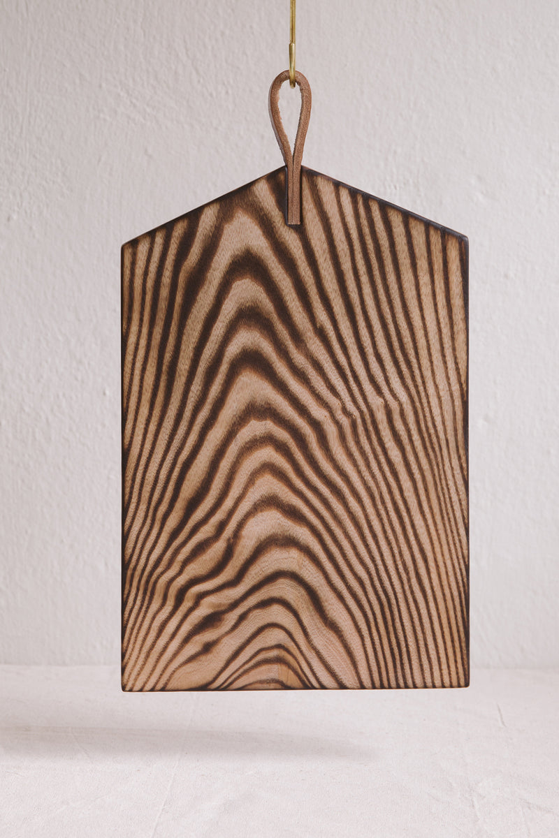 Handmade by Ambrose Vevers. These chopping boards are made from ash wood, which has been scorched with a naked flame and finished with food safe oil. A leather loop is at the top of the board for easy display and storage. The shape of this board is a rectangle with a slanted edges at the top like a house.